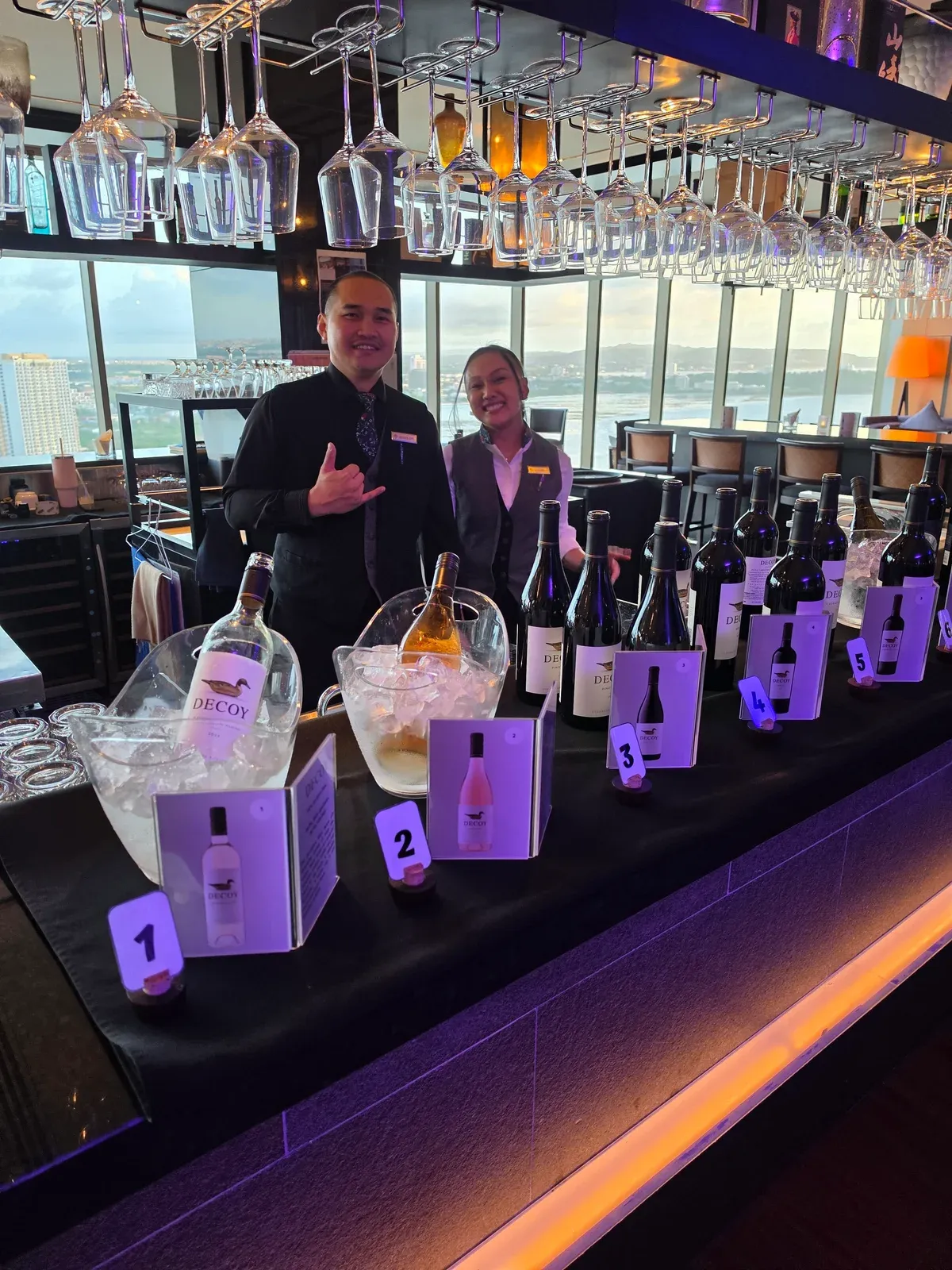 Two staff members smiling behind the bar at Decoy Uncorked, with the numbered line-up of Decoy bottles and ice buckets in front of them