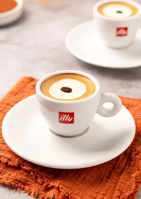 An illy espresso cup with a milk foam circle and a single coffee bean, on a white saucer over an orange napkin